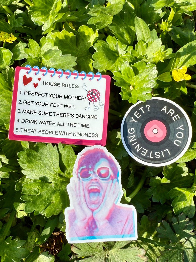 May include: A collection of stickers on a bed of green foliage. One sticker is a notepad with "HOUSE RULES" and bullet points. Another is a circular sticker with a record design and the words "ARE YOU LISTENING YET?". The third sticker is a colorful image of a person.