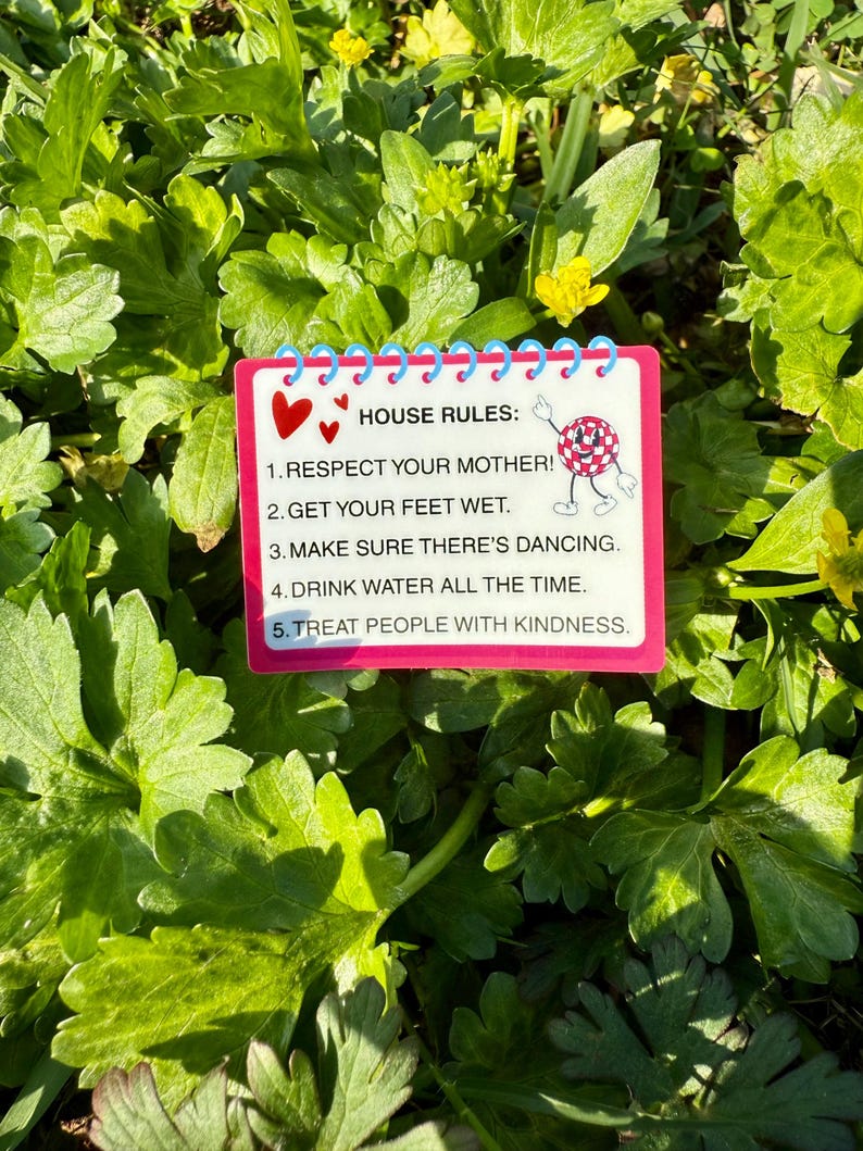 May include: A rectangular sign with a pink border and blue spiral binding, featuring the text "HOUSE RULES:" followed by five rules. The sign is decorated with red hearts and a cartoon character. The background is lush green foliage.