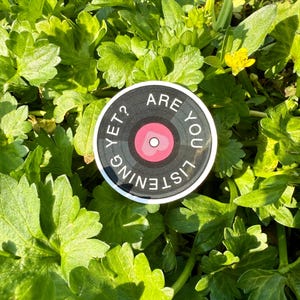 May include: A round sticker with a vinyl record design. The sticker has a black background with white text that reads "ARE YOU LISTENING YET?". The center of the record is pink with a white heart. The sticker is set against a background of green leaves.