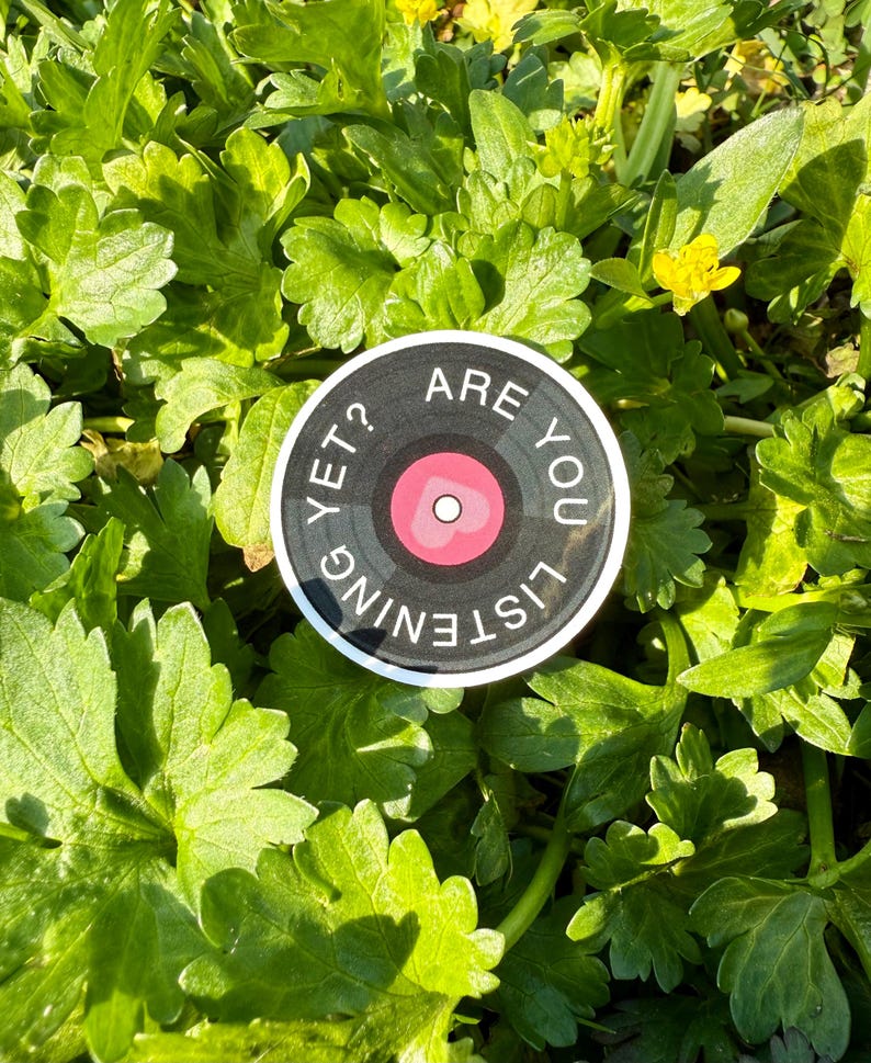 May include: A round sticker with a vinyl record design. The sticker has a black background with white text that reads "ARE YOU LISTENING YET?". The center of the record is pink with a white heart. The sticker is set against a background of green leaves.