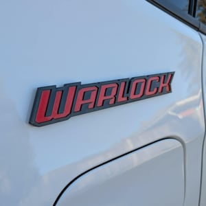 Op de afbeelding: Een close-up van het zijpaneel van een wit voertuig met een rood en zwart "WARLOCK" embleem. Het embleem is rechthoekig met een zwarte omtrek en rode letters. Het oppervlak van het voertuig is schoon en reflecterend.