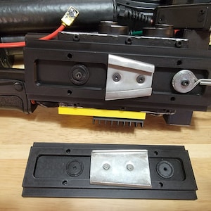 Op de afbeelding: Een zwart kunststof onderdeel met een metalen plaat en een kleine haak. Het onderdeel heeft meerdere gaten en een stukje gele tape.