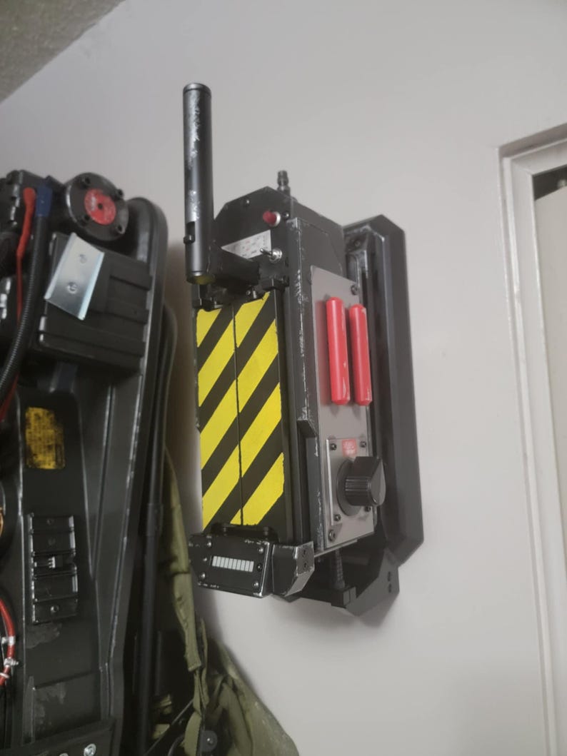 May include: A black and yellow Ghostbusters proton pack replica with a red button and a black antenna. The pack is mounted on a black wall mount.