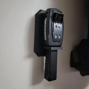 May include: A black wall mount for a handheld device, with a textured handle and a rectangular display area. The mount is made of plastic and has a simple design.
