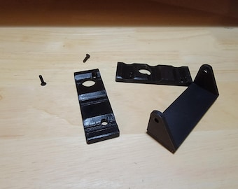 Puede incluir: Componentes negros impresos en 3D, incluidos soportes y tornillos, están dispuestos sobre una superficie de madera clara. Los componentes tienen agujeros para tornillos y una superficie texturizada. La imagen muestra las piezas de un sistema de montaje.
