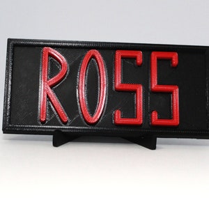 May include: A black rectangular sign with the name "ROSS" in red, raised letters. The sign is supported by a small black stand. The letters are bold and stand out against the black background.