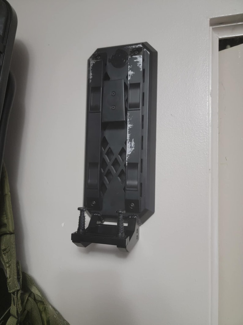 May include: A black wall mount with a textured surface and a small shelf. The mount has two screws on the bottom and a rectangular opening on the top.