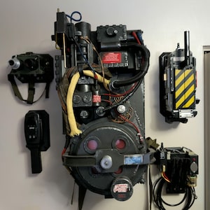 Può includere: Una replica dettagliata di un pacco protonico Ghostbusters e delle relative attrezzature, montata su un muro. Il pacco è principalmente nero e grigio, con accenti rossi, gialli e blu. Componenti aggiuntivi includono una trappola, un misuratore PKE e una trappola per fantasmi.