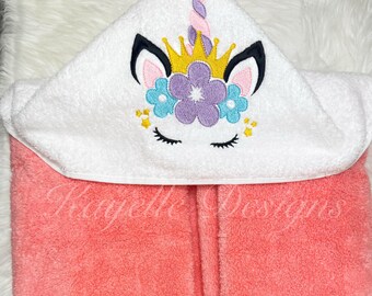 May include: A pink hooded towel with a white unicorn embroidered on the hood. The unicorn has a gold crown, purple flowers, and closed eyes.