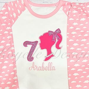May include: Pink and white pajamas with a white cloud pattern. The top has a pink and white glitter number 7, a pink silhouette of a Barbie doll, and the name "Arabella" embroidered in pink.