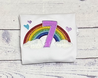 May include: A white t-shirt with a rainbow and a purple number 7 embroidered on it. There are also small blue and purple hearts embroidered around the rainbow.