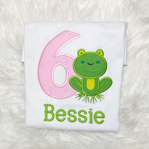 May include: A white t-shirt featuring an embroidered design of a large pink number six next to a green frog with a yellow belly. Below the frog, the name Bessie is embroidered in green letters. The design includes the text Rayelle Designs.