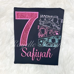 May include: Black t-shirt with a large, glittery pink number 7. The shirt features embroidered text that reads "Safiyah" in pink script, and a comic-style graphic with the text "HUNTRX", "MIRA", "RUMI", and "ZOEY".
