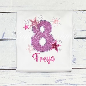 Peut inclure: T-shirt blanc avec un grand chiffre 8 violet pailleté et le nom "Freya" en rose. Des étoiles et des volutes roses et pailletées entourent le chiffre, créant un design festif.