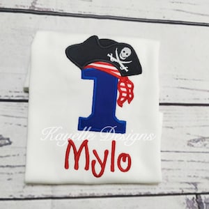 May include: White onesie with a blue number one and a red and white pirate hat with a skull and crossbones. The name "Mylo" is embroidered in red below the number.
