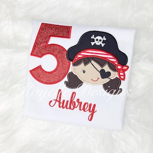 Pode incluir: Número 5 de glitter vermelho com um chapéu de pirata listrado de vermelho e branco com uma caveira e ossos cruzados. O pirata está usando um tapa-olho e tem cabelo castanho. O nome Aubrey está bordado em vermelho abaixo do número.