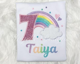 May include: A white t-shirt with a pink glittery number 7, a rainbow with a white cloud, and the name "Taiya" embroidered in rainbow colors.