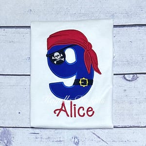 May include: White t-shirt with a blue number nine, a red pirate hat, and a skull and crossbones eye patch. The name "Alice" is embroidered below the number.