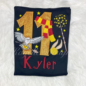 Könnte beinhalten: Schwarzes T-Shirt mit goldglitzernder "11" und rotem Namen. Mit Eule, Schal, Zauberstab und goldenem Schnatz. Gelbe Sterne sind um das Design verstreut. Das Design ist gestickt.