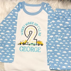 May include: A two-piece pajama set. The white top has blue sleeves with a cloud pattern and a blue neckline. The front of the top has the text "WHEN I WAKE UP I'LL BE 2 GEORGE" with construction vehicles. The matching pants are light blue with a cloud pattern.