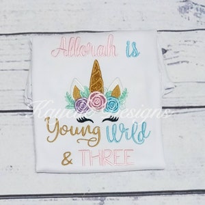 May include: A white t-shirt with a unicorn design and the text "Allorah is Young Wild & Three". The unicorn has a gold horn, pink and blue flowers, and gold glitter eyelashes.