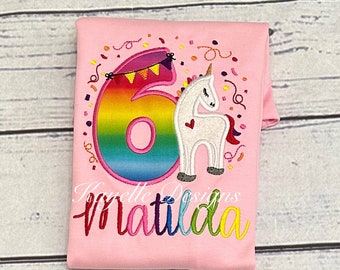 May include: Pink t-shirt with a rainbow number six and a white unicorn with a pink heart. The shirt says "Matilda" in rainbow letters.