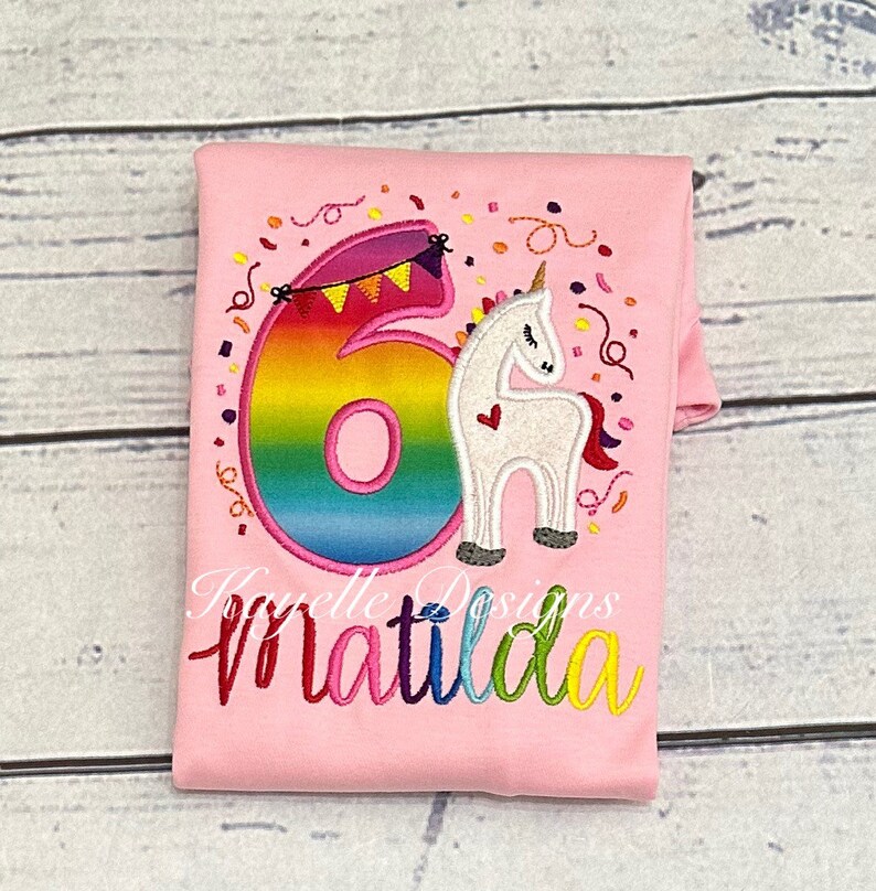 May include: Pink t-shirt with a rainbow number six and a white unicorn with a pink heart. The shirt says "Matilda" in rainbow letters.