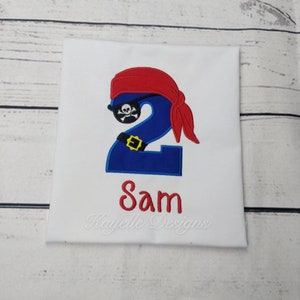 May include: A white t-shirt with a blue number two wearing a red pirate bandana and an eye patch. The number two has a gold belt. The name Sam is embroidered below the number. The text "Kayelle Designs" is embroidered below the name.