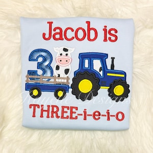 May include: Light blue shirt with a tractor and cow design. The shirt features the text "Jacob is 3 THREE-i-e-i-o" in red embroidery. The tractor is blue with yellow wheels, and the cow is black and white.