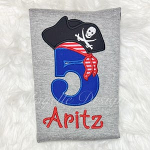 May include: A gray t-shirt with a pirate-themed design. The design includes a black pirate hat with a skull and crossbones, a blue number 5, and the name "Aritz" in red. The pirate hat has a red and white striped bandana.