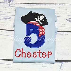 May include: Pale blue t-shirt featuring a pirate design. It has a black pirate hat with a skull and crossbones, a large blue number 5, and the name "Chester" in red. This is likely a birthday shirt for a child.
