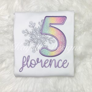 Może przedstawiać: Biała koszulka z błyszczącą liczbą 5 w kolorze tęczy, srebrną śnieżynką i imieniem "Florence" wyszytym na fioletowo.