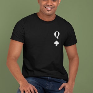 May include: Black t-shirt with a white Queen of Spades design. The shirt features a small graphic of the letter "Q" above a spade symbol. The model is wearing blue jeans and is seated on a wooden stool.