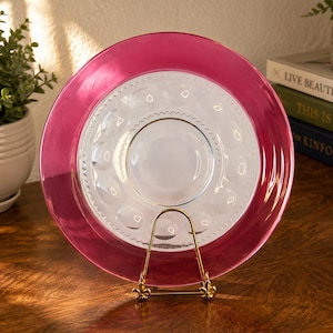 May include: A round glass plate featuring a wide, translucent magenta rim and a clear center with a textured pattern of circular indentations. The plate is displayed upright on a gold-toned metal stand, showcasing its decorative design.