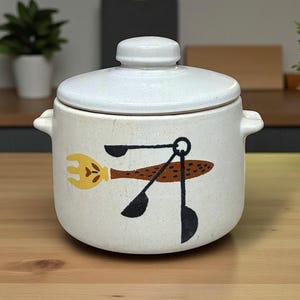 May include: A white ceramic lidded pot with a speckled finish and two side handles. The front features a graphic illustration of a yellow fork and a set of black measuring spoons arranged in a cluster, centered on the body of the round container.