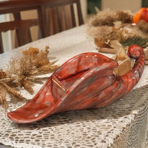 May include: A decorative, red and gray ceramic swan-shaped bowl. The bowl has a curved, elongated design with a textured surface. The swan's head is raised, and the bowl sits on a table with a lace tablecloth. The bowl is a decorative item.