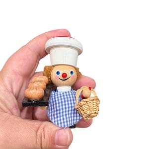 May include: A wooden figurine of a baker wearing a white hat with a red trim, a blue and white checkered apron, and holding a basket of bread. The baker has a red nose and blue eyes.