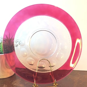 May include: A clear glass plate with a vibrant pink rim. The plate has a white center with a decorative pattern and is displayed on a gold-colored stand. A small potted plant is visible in the background.