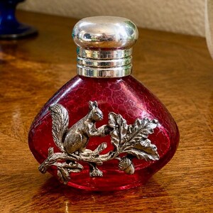 May include: A small, rounded perfume bottle made of deep red crackle glass with a silver-toned metal cap. The front of the bottle is decorated with a detailed silver-toned metal applique featuring a squirrel perched on an oak branch with leaves.