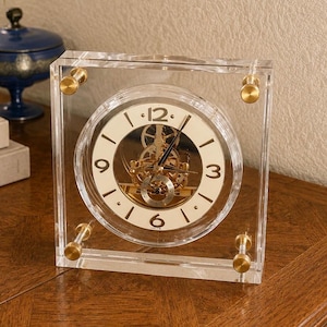May include: A square clear acrylic table clock featuring a round white dial with black Arabic numerals and visible gold-toned mechanical gears. The clock is held together by four gold-toned metal bolts at each corner, showcasing a transparent design.