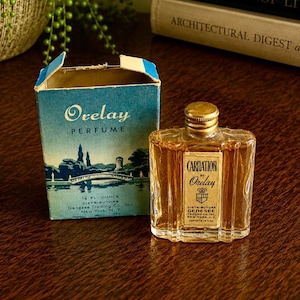 May include: A vintage Orelay Carnation perfume bottle with a gold cap and a rectangular glass body, accompanied by its original blue cardboard box featuring a landscape illustration and the text Orelay Perfume, 1/4 fl. ounce, Genesee Trading Co.