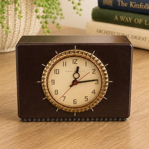 May include: A rectangular dark brown General Electric clock with a cream-colored circular face, black numerals, and a metallic gold-toned gear-shaped bezel. The clock features black hour and minute hands, a thin red second hand, and radial markers.
