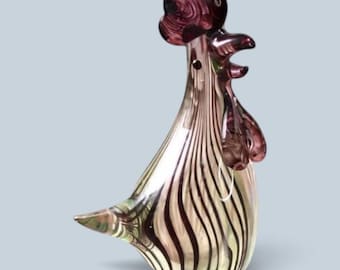 Op de afbeelding: Een glazen haanfiguur met een doorschijnend lichaam voorzien van verticale donkerpaarse en heldere strepen. Het beeldje heeft een dieppaarse kam en lellen, een puntige staart en een gladde, reflecterende afwerking over het gehele oppervlak.