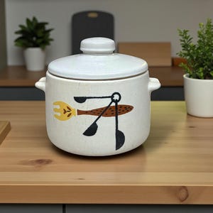 May include: A white ceramic casserole dish with a lid, featuring a whimsical design of a fork and spoons in brown, yellow, and black. The dish has two small handles and is suitable for kitchen use.