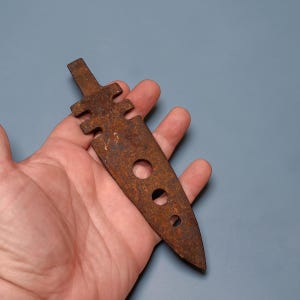 May include: A rusty, metal arrowhead with a flat, elongated body and a notched handle. Three circular holes are drilled into the blade. The arrowhead is held in a hand against a blue-gray background.