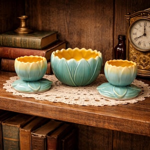 May include: Three ceramic bowls with a lotus flower design. The bowls are in shades of blue and yellow. The largest bowl sits in the center, with two smaller bowls on either side. The bowls are on a lace doily, with a wooden shelf and antique clock in the background.