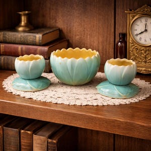 May include: Three ceramic bowls with a floral design. The bowls are light blue with a yellow interior. Two smaller bowls sit on leaf-shaped bases. The largest bowl is in the center. The bowls are on a lace doily on a wooden shelf.