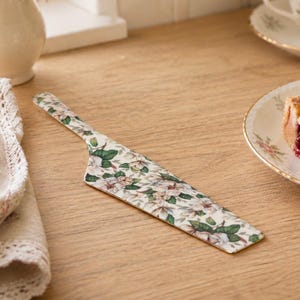May include: A floral-patterned cake server with a cream background and a gold edge. The server features a design of white and pink flowers with green leaves. The server is on a wooden surface.