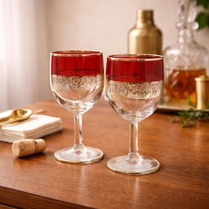 May include: Two clear wine glasses with a red rim and a gold filigree design. The glasses have a gold rim at the base. The glasses are on a wooden table. A cork, napkins, and gold spoons are also on the table.