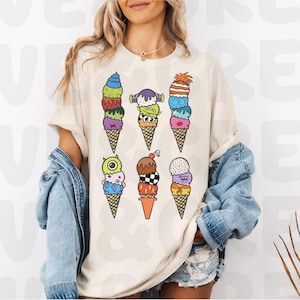 May include: Cream-colored t-shirt featuring a colorful graphic of six monster-themed ice cream cones. Each cone has a unique design with various colors and monster-like features. The shirt is worn by a person with blonde hair and a denim jacket.